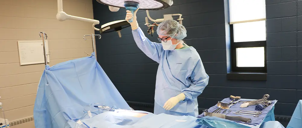 Healthcare student in a surgical gown, mask, gloves, and protective eyewear adjusts an operating room light beside a prepared surgical table and sterilized instruments.